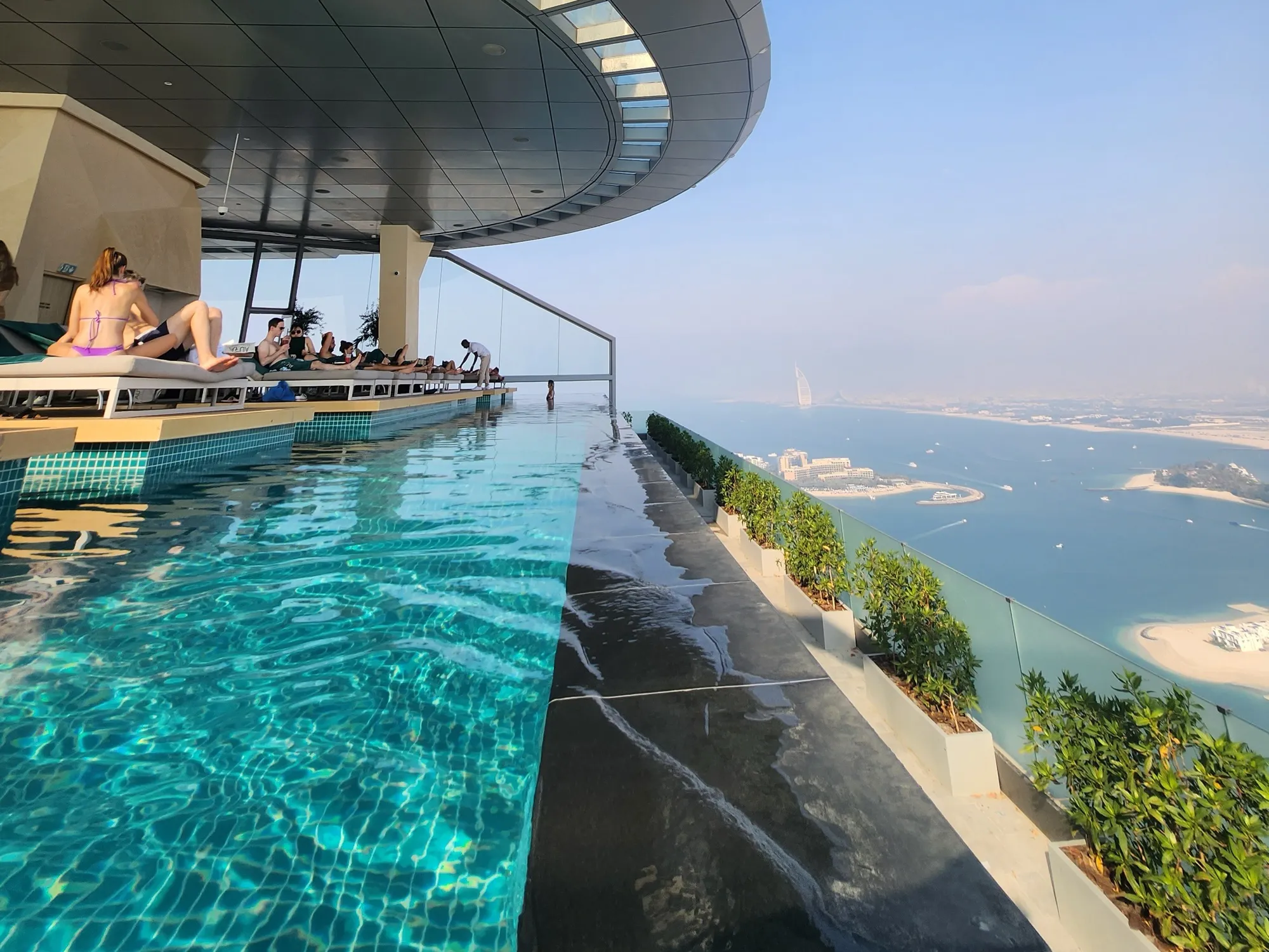 Guests on loungers beside the turquoise infinity pool at AURA Skypool on The Palm Tower, with Palm Jumeirah and the Burj Al Arab in the haze beyond