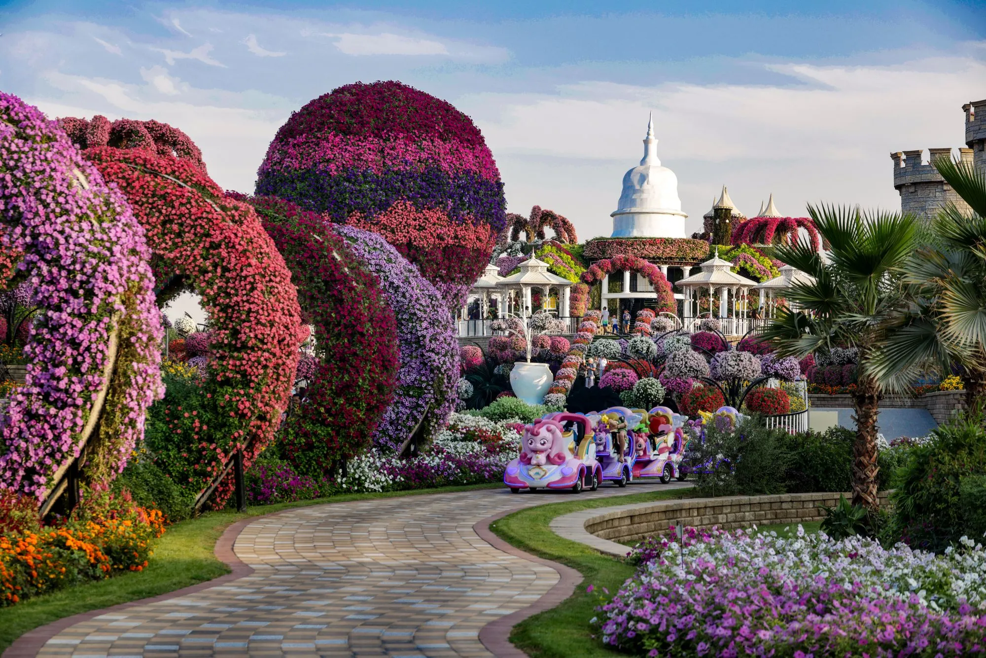 Tunnels of pink and purple petunias arching over a paved path at Dubai Miracle Garden, with the floral castle in the distance