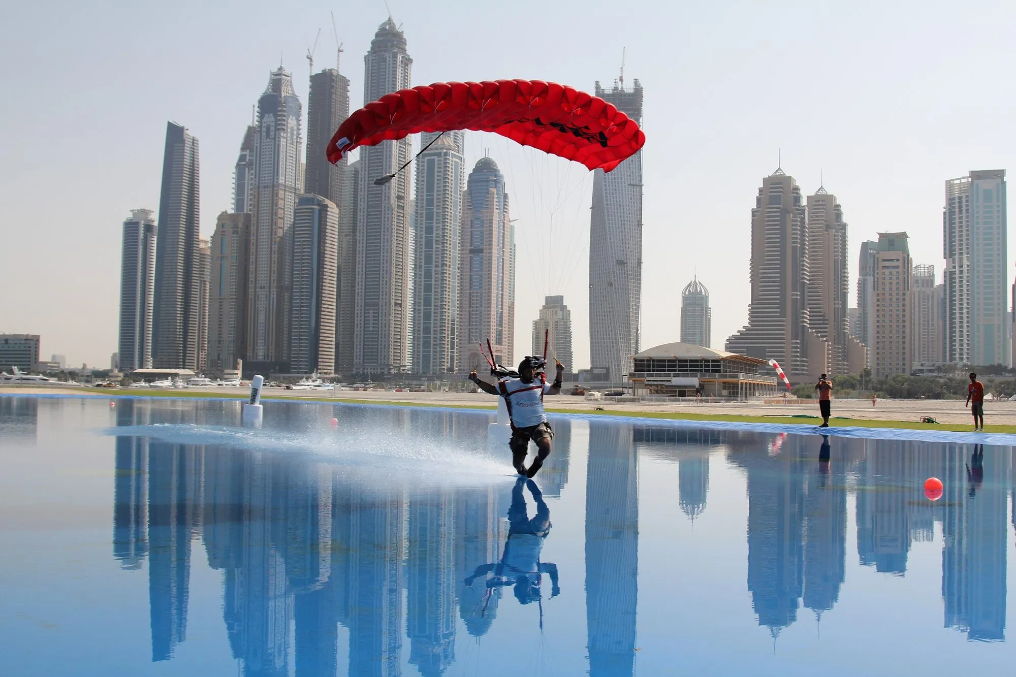 A skydiver landing a red canopy across the swoop pond at Skydive Dubai with the Dubai Marina skyline reflected in the water