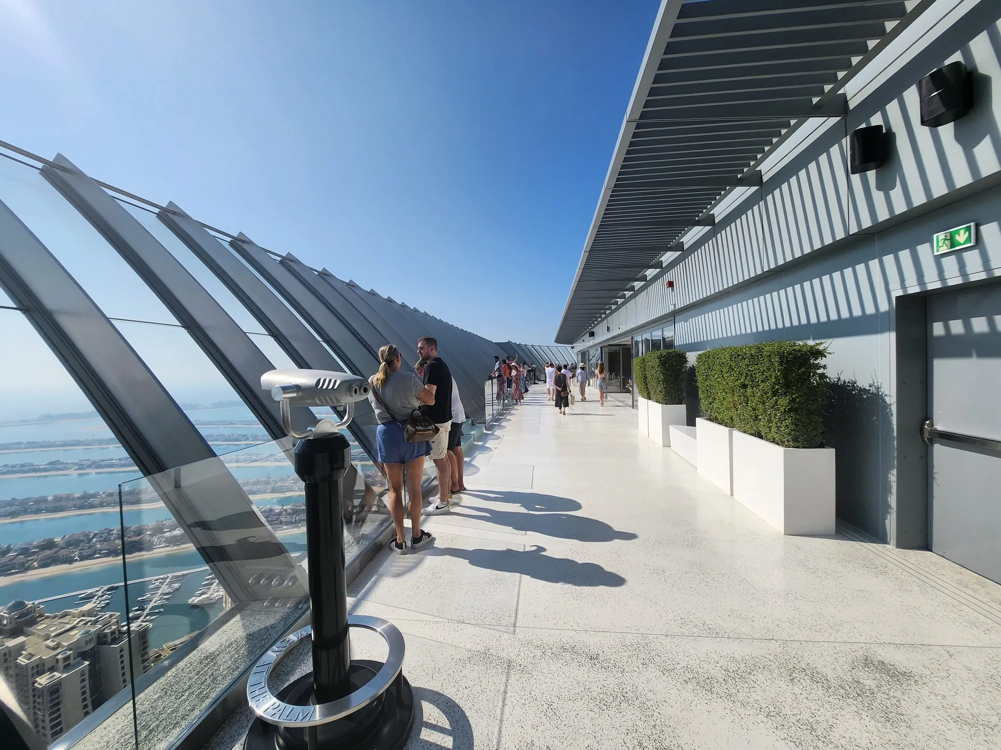 The sunlit glass-walled observation deck of The View at The Palm with visitors looking out over Palm Jumeirah's fronds